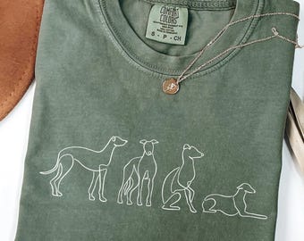 May include: Olive green t-shirt with a white line drawing of four dogs in various poses. The shirt has a crew neck and a small tag on the collar. A gold necklace with a pendant is draped across the shirt.