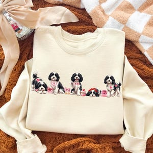 May include: Cream-colored sweatshirt featuring a line of five black and white puppies with pink heart accents and gifts. The puppies are in various poses, with some sitting and others lying down. The sweatshirt has long sleeves.
