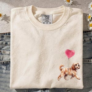 Op de afbeelding: Een crèmekleurig T-shirt met ronde hals en een Comfort Colors label. Het shirt heeft een afbeelding van een bruine hond die loopt terwijl hij een roze hartvormig ballonnetje aan een touwtje vasthoudt. Het T-shirt ligt op een blauwe spijkerbroek.