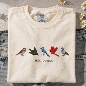 Op de afbeelding: Een crèmekleurig T-shirt met ronde hals en een afbeelding van vijf verschillende vogels op een rij boven de tekst SEED DEALER. Het shirt heeft een Comfort Colors-merklabel in de nek en ligt op een blauwe spijkerbroek van denim.