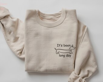 Könnte beinhalten: Ein beiger Pullover mit einer schwarzen Dackel-Illustration und dem Text "It's been a long day".