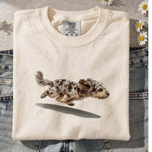 May include: Cream-colored t-shirt featuring a watercolor illustration of a running dog with brown and gray spots. The shirt is folded on top of a pair of blue jeans. The tag reads "Comfort Colors".