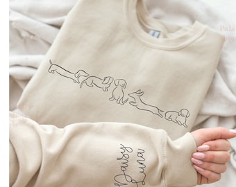 Puede incluir: Una sudadera beige con un dibujo lineal de cuatro dachshunds en la parte delantera. La manga tiene el texto "Daisy Luna" escrito en cursiva.