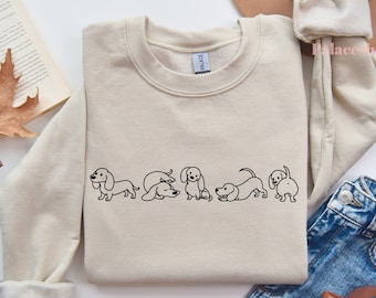 Könnte beinhalten: Ein beiger Pullover mit einer schwarzen Umrandung von fünf Dackeln in verschiedenen Posen. Die Hunde sind alle in die gleiche Richtung ausgerichtet und in einer Reihe angeordnet.