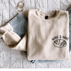 Può includere: Una felpa beige con una grafica nera che dice "Wake & Bake" con un'illustrazione di un cartone animato di una pagnotta di pane. La felpa è piegata e appoggiata su una superficie testurizzata grigio chiaro con un paio di orecchini a cerchio dorati.