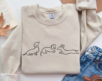 Könnte beinhalten: Ein beiger Pullover mit einer einfachen Linienzeichnung von drei Dackeln auf der Vorderseite. Die Hunde sind in verschiedenen Posen, einer sitzt, einer liegt und einer rennt.