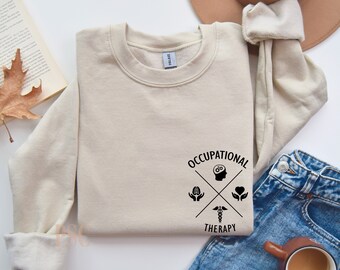 Puede incluir: Una sudadera beige con un diseño gráfico negro que dice "Occupational Therapy" con una cruz y símbolos que representan el cerebro, las manos y un corazón.