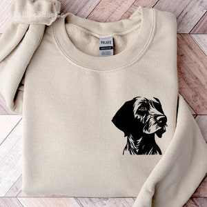 May include: A beige sweatshirt with a black silhouette of a dog's head on the left side of the chest. The sweatshirt is folded and laid flat on a wooden surface.
