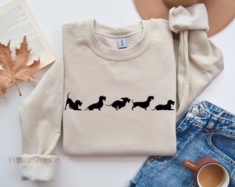 Könnte beinhalten: Ein beiger Pullover mit einer schwarzen Silhouette von fünf Dackeln, die auf der Vorderseite aufgedruckt sind. Die Hunde stehen in einer Reihe, schauen nach rechts und sind in verschiedenen Posen.