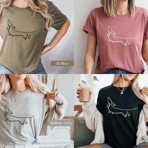 May include: Four different color t-shirts featuring a simple line drawing of a dachshund. The colors are olive, mauve, athletic grey, and black.