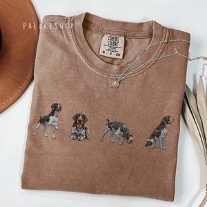 Könnte beinhalten: Ein braunes Comfort Colors T-Shirt mit vier Hundeillustrationen. Eine silberne Halskette mit Anhänger liegt auf dem Shirt. Die Hunde sind in verschiedenen Posen. Das Shirt hat einen Rundhalsausschnitt und kurze Ärmel.