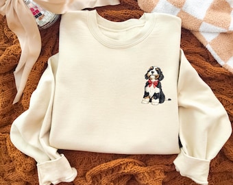 Puede incluir: Sudadera color crema con un cachorro Bernedoodle bordado con una pajarita roja. La parte superior de manga larga tiene cuello redondo y puños acanalados. El cachorro es negro, blanco y marrón.