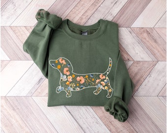May include: A green sweatshirt with a white outline of a dachshund filled with a floral pattern. The flowers are in shades of pink, yellow, and blue.