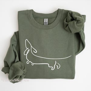 May include: A green sweatshirt with a white outline of a dachshund dog. The sweatshirt has a crew neck and long sleeves.