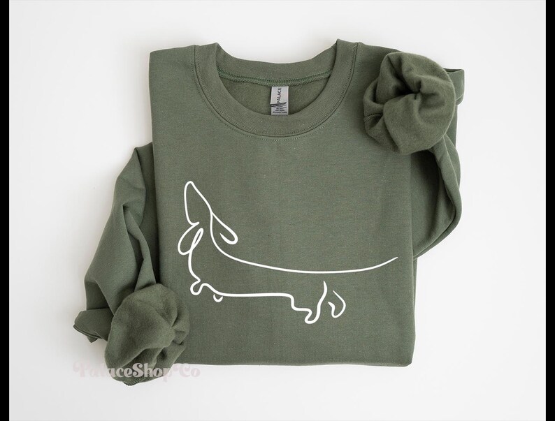 May include: A green sweatshirt with a white outline of a dachshund dog. The sweatshirt has a crew neck and long sleeves.