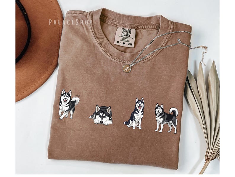 Pu&ograve; includere: Una maglietta marrone Comfort Colors con quattro disegni di cani husky ricamati in bianco e nero. La maglietta ha un girocollo e maniche corte. Una collana d'argento con un ciondolo &egrave; drappeggiata sulla maglietta. La parola "PALACESHOP" &egrave; stampata in alto a sinistra.