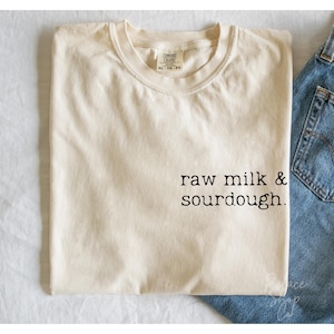 Può includere: Una maglietta color crema con la scritta "raw milk & sourdough." stampata in nero sul davanti. La maglietta è appoggiata su una superficie bianca con un paio di jeans blu sullo sfondo.