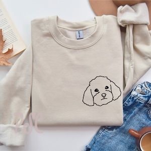 Op de afbeelding: Een beige sweatshirt met een zwarte omtrek van een poedels gezicht. De sweatshirt heeft een ronde hals en lange mouwen. De afbeelding staat op een wit oppervlak met een boek en een herfstblad.