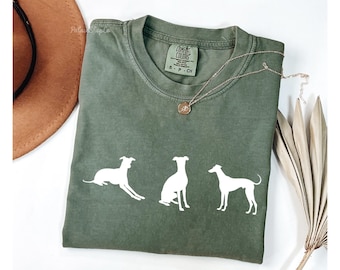 May include: Olive green t-shirt with three white dog silhouettes. The shirt has a crew neck and a small tag on the collar. A gold necklace with a pendant is draped across the shirt. The shirt is folded on a white surface.