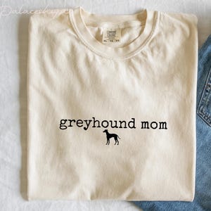 May include: Cream-colored t-shirt with the text "greyhound mom" in black, featuring a silhouette of a greyhound dog. The shirt is folded, with a pair of blue jeans visible on the right side.
