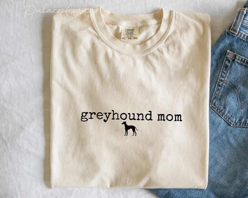 May include: Cream-colored t-shirt with the text "greyhound mom" in black, featuring a silhouette of a greyhound dog. The shirt is folded, with a pair of blue jeans visible on the right side.
