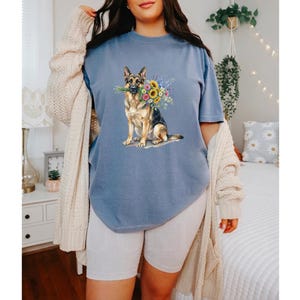 Peut inclure: T-shirt bleu avec une illustration &agrave; l'aquarelle d'un berger allemand tenant un bouquet de tournesols et d'autres fleurs color&eacute;es. Le chien est repr&eacute;sent&eacute; de mani&egrave;re r&eacute;aliste, avec des d&eacute;tails sur la fourrure et une expression douce. Le t-shirt est de style d&eacute;contract&eacute;.