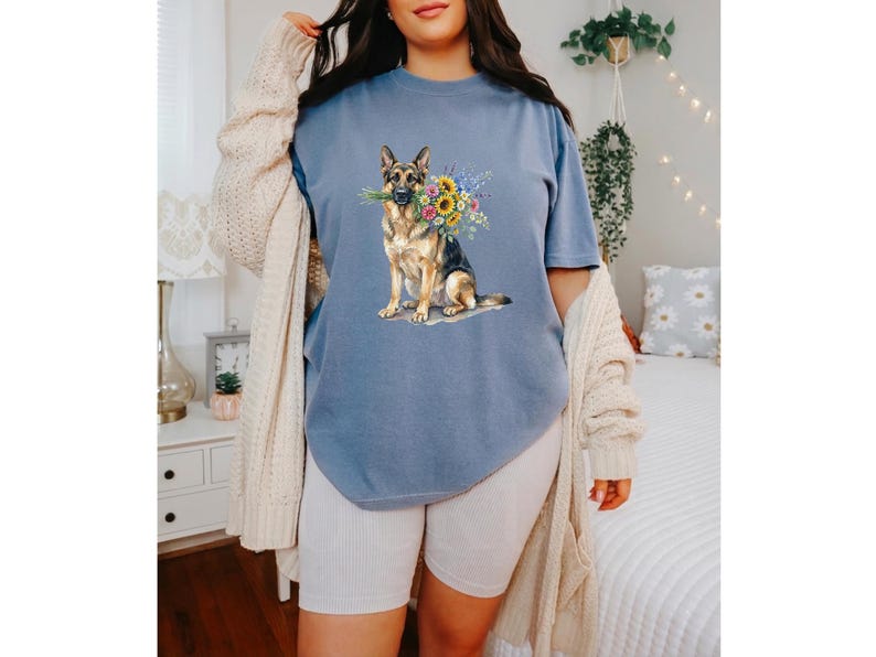 Peut inclure: T-shirt bleu avec une illustration &agrave; l'aquarelle d'un berger allemand tenant un bouquet de tournesols et d'autres fleurs color&eacute;es. Le chien est repr&eacute;sent&eacute; de mani&egrave;re r&eacute;aliste, avec des d&eacute;tails sur la fourrure et une expression douce. Le t-shirt est de style d&eacute;contract&eacute;.