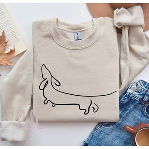 May include: A beige sweatshirt with a simple line drawing of a dachshund on the front.