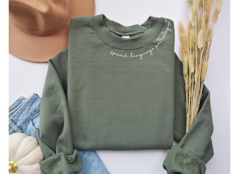 Puede incluir: Una sudadera verde con el texto "speech language pathologist" bordado en blanco en el cuello.