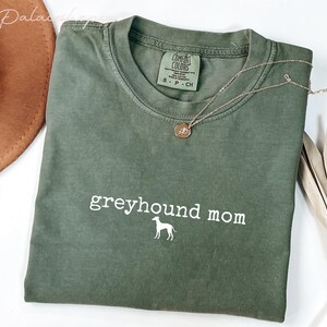 May include: An olive green t-shirt with the words "greyhound mom" and a greyhound silhouette printed in white. A gold necklace with a pendant rests on the shirt. A brown hat and dried plant are in the background.