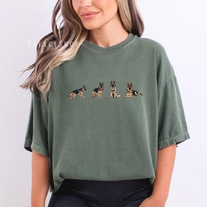 Op de afbeelding: Olijfgroen T-shirt met een geborduurd ontwerp van een Duitse herder in vier verschillende poses. Het shirt heeft een relaxte pasvorm en een ronde hals. De tekst "PALACEWOOD" is zichtbaar linksboven op het shirt.