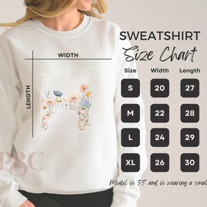 Puede incluir: Sudadera blanca con un dise&ntilde;o floral y una tabla de tallas. La tabla muestra las tallas S, M, L y XL con las medidas de ancho y largo correspondientes en pulgadas. La modelo mide 5'5" y lleva una talla peque&ntilde;a.