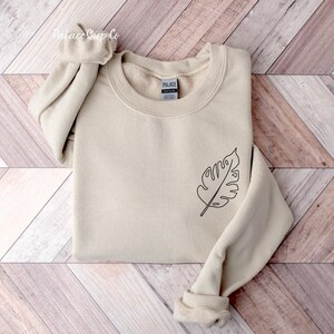 May include: A beige sweatshirt with a simple line drawing of a leaf on the left side of the chest. The sweatshirt has a crew neck and long sleeves.