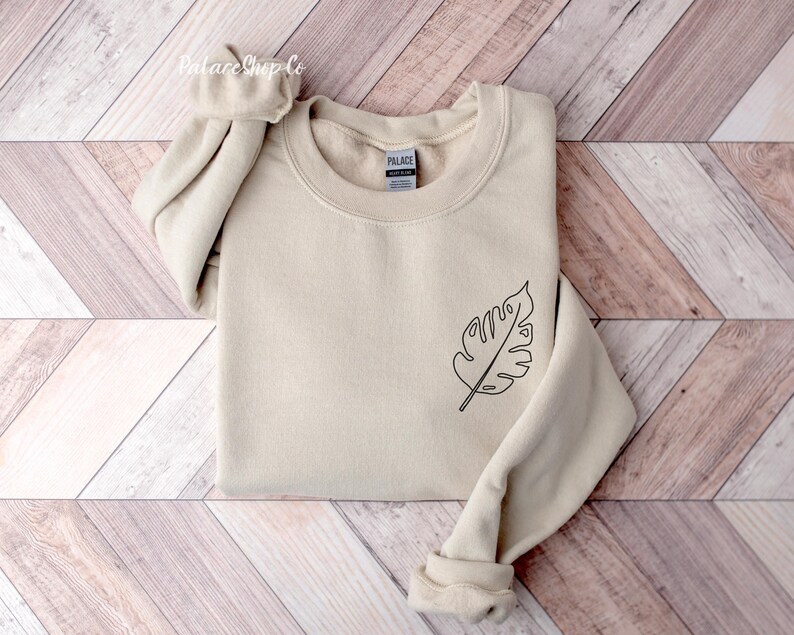 May include: A beige sweatshirt with a simple line drawing of a leaf on the left side of the chest. The sweatshirt has a crew neck and long sleeves.