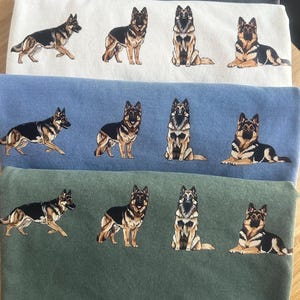 Op de afbeelding: Drie stapels stof in wit, blauw en groen, elk met geborduurde ontwerpen van Duitse herdershonden. De honden worden afgebeeld in verschillende poses, waaronder staan, zitten en liggen, met gedetailleerde zwarte, bruine en bruine markeringen.