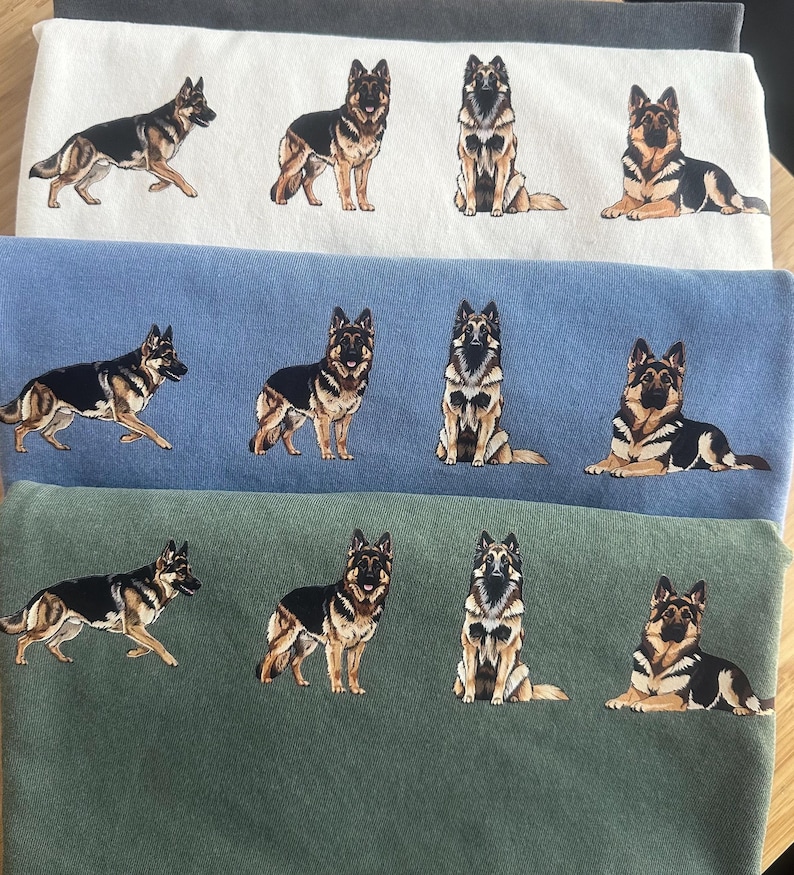 Op de afbeelding: Drie stapels stof in wit, blauw en groen, elk met geborduurde ontwerpen van Duitse herdershonden. De honden worden afgebeeld in verschillende poses, waaronder staan, zitten en liggen, met gedetailleerde zwarte, bruine en bruine markeringen.