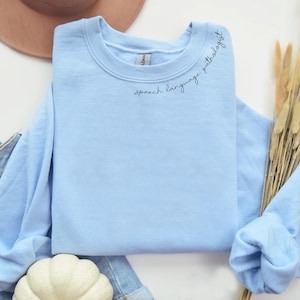 Puede incluir: Una sudadera de color azul claro con el texto "speech language pathologist" bordado en el cuello.