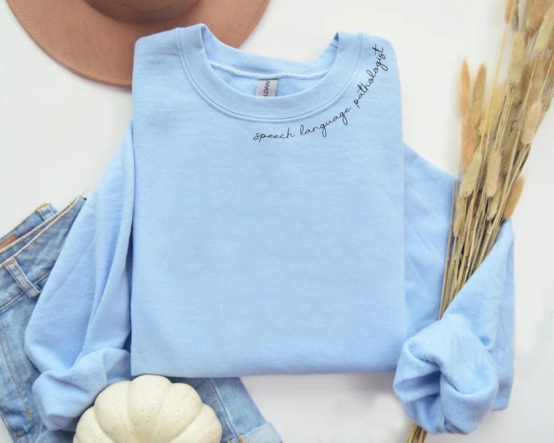 Puede incluir: Una sudadera de color azul claro con el texto "speech language pathologist" bordado en el cuello.