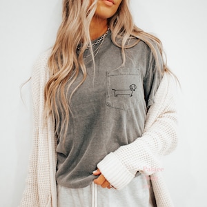 May include: A gray t-shirt with a pocket featuring a simple black line drawing of a dachshund. The shirt is worn by a person with long blonde hair and a white cardigan.