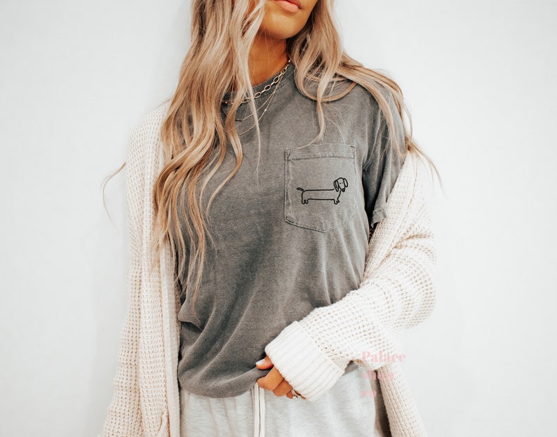 May include: A gray t-shirt with a pocket featuring a simple black line drawing of a dachshund. The shirt is worn by a person with long blonde hair and a white cardigan.