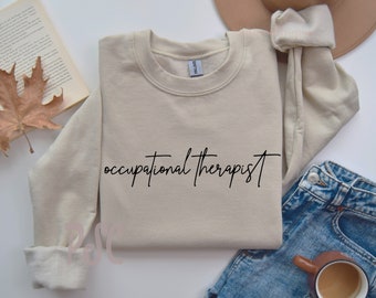 Puede incluir: Una sudadera beige con el texto "occupational therapist" escrito en cursiva negra. La sudadera está doblada y colocada sobre una superficie blanca con un par de pantalones vaqueros azules y una taza de café.