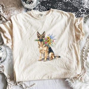 Peut inclure: T-shirt cr&egrave;me avec une illustration &agrave; l'aquarelle d'un berger allemand tenant un bouquet de tournesols et d'autres fleurs color&eacute;es dans sa gueule. Le chien est assis droit. Le t-shirt a un col rond.