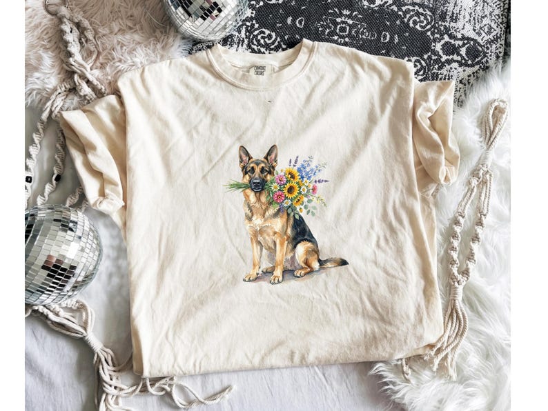 Peut inclure: T-shirt cr&egrave;me avec une illustration &agrave; l'aquarelle d'un berger allemand tenant un bouquet de tournesols et d'autres fleurs color&eacute;es dans sa gueule. Le chien est assis droit. Le t-shirt a un col rond.