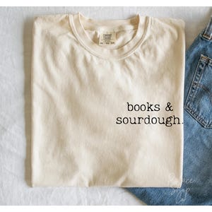 Può includere: Una maglietta color crema con la scritta "books & sourdough." stampata in nero sul davanti.