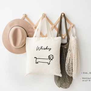 May include: A white canvas tote bag with black text that says "Whiskey" and a black line drawing of a dachshund. The bag is hanging on a wooden wall rack with two other bags.