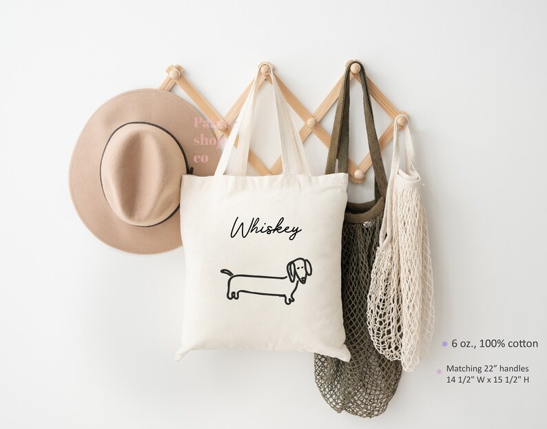 May include: A white canvas tote bag with black text that says "Whiskey" and a black line drawing of a dachshund. The bag is hanging on a wooden wall rack with two other bags.
