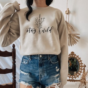 May include: A beige sweatshirt with the text "stay wild" and a floral graphic printed on the front. The sweatshirt is being worn by a person wearing denim shorts.