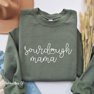 Può includere: Una felpa verde con la scritta "sourdough mama" in lettere bianche. Sopra la parola "mama" c'è una piccola illustrazione di una pagnotta di pane a lievitazione naturale.
