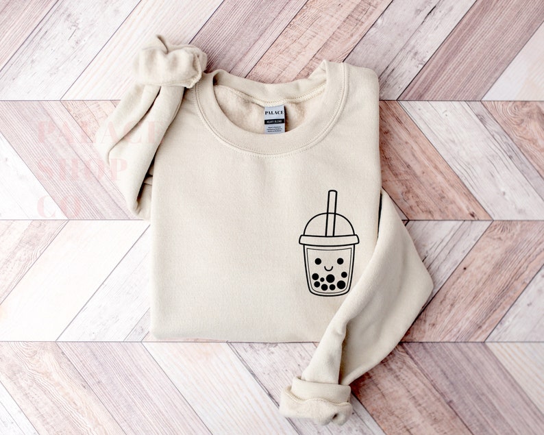 May include: A beige sweatshirt with a black graphic of a smiling boba tea cup with black boba pearls. The sweatshirt has a crew neck and long sleeves.