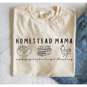 Può includere: Una maglietta beige con testo nero che recita "Homestead Mama making. schooling. steadying" con illustrazioni di una pagnotta di pane, una pila di libri e una gallina.